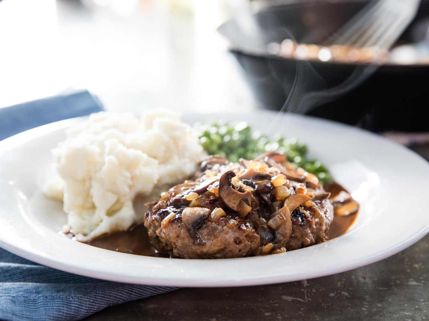 A finished serving of Salisbury steak, served alongside mashed potatoes and green peas.