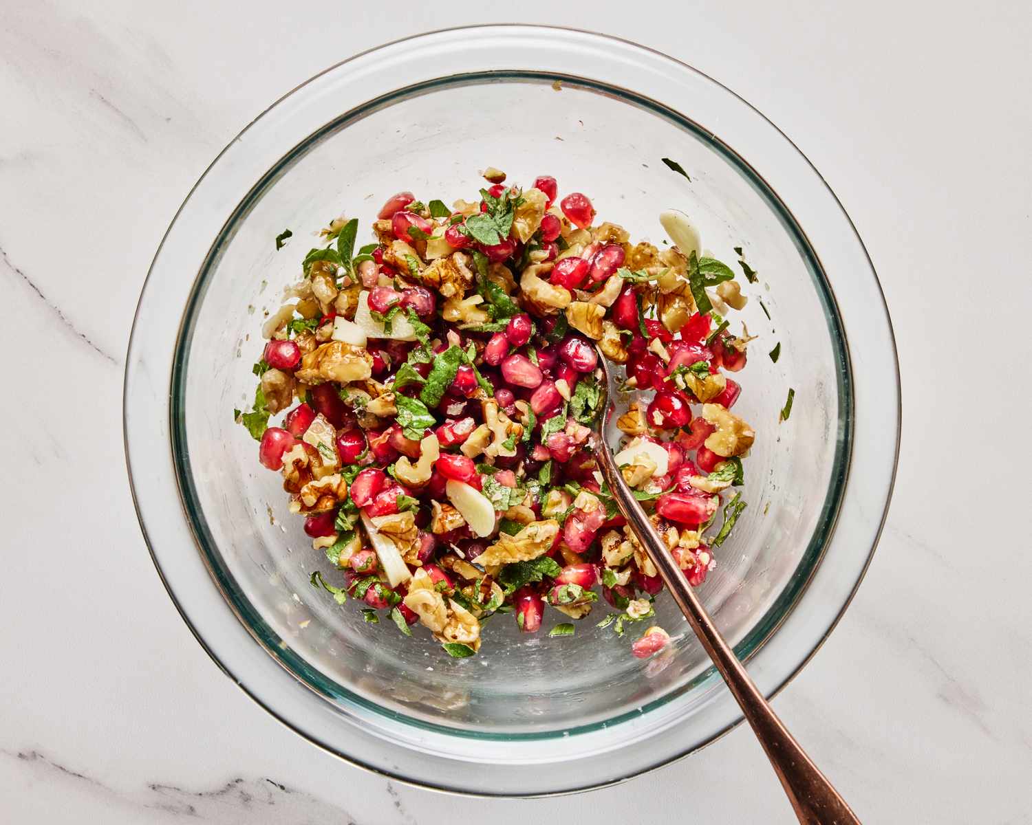 A glass bowl containing a mixture of pomegranate seeds chopped nuts herbs and other ingredients with a spoon inside