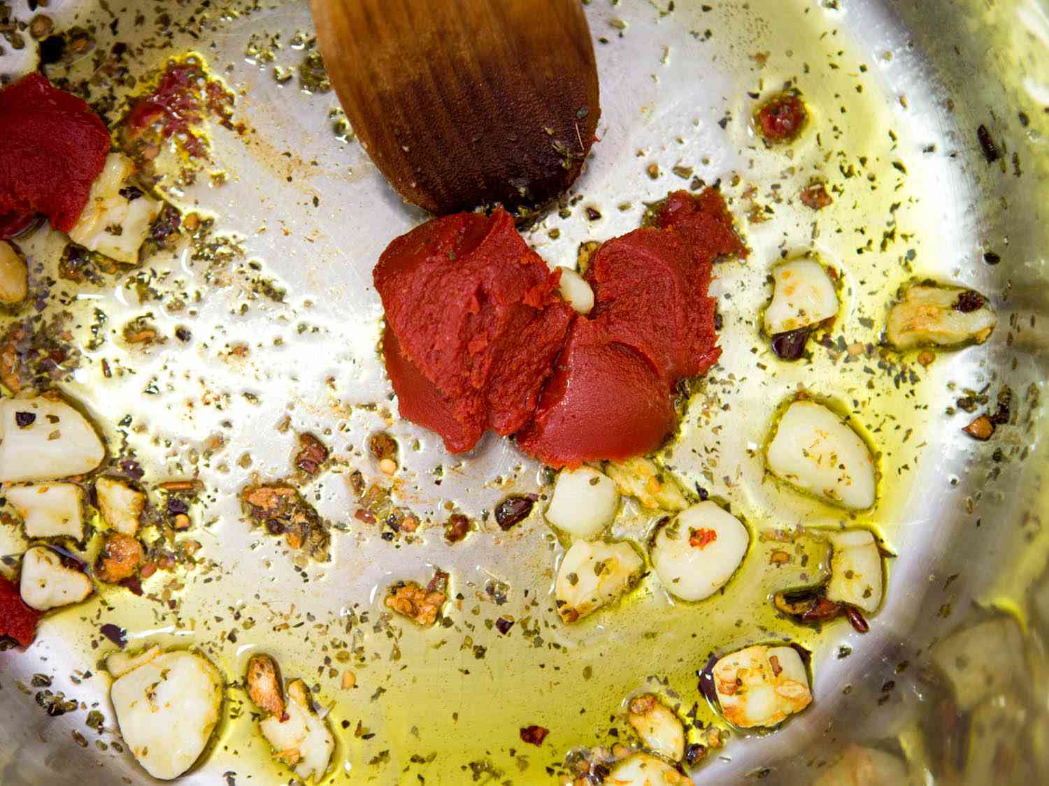 Adding tomato paste to pot with garlic, oregano, red pepper flakes, and olive oil.