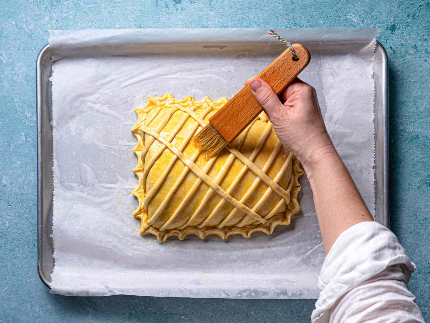 A hand brushing an egg wash onto a latticetopped pastry on parchment paper on a baking sheet
