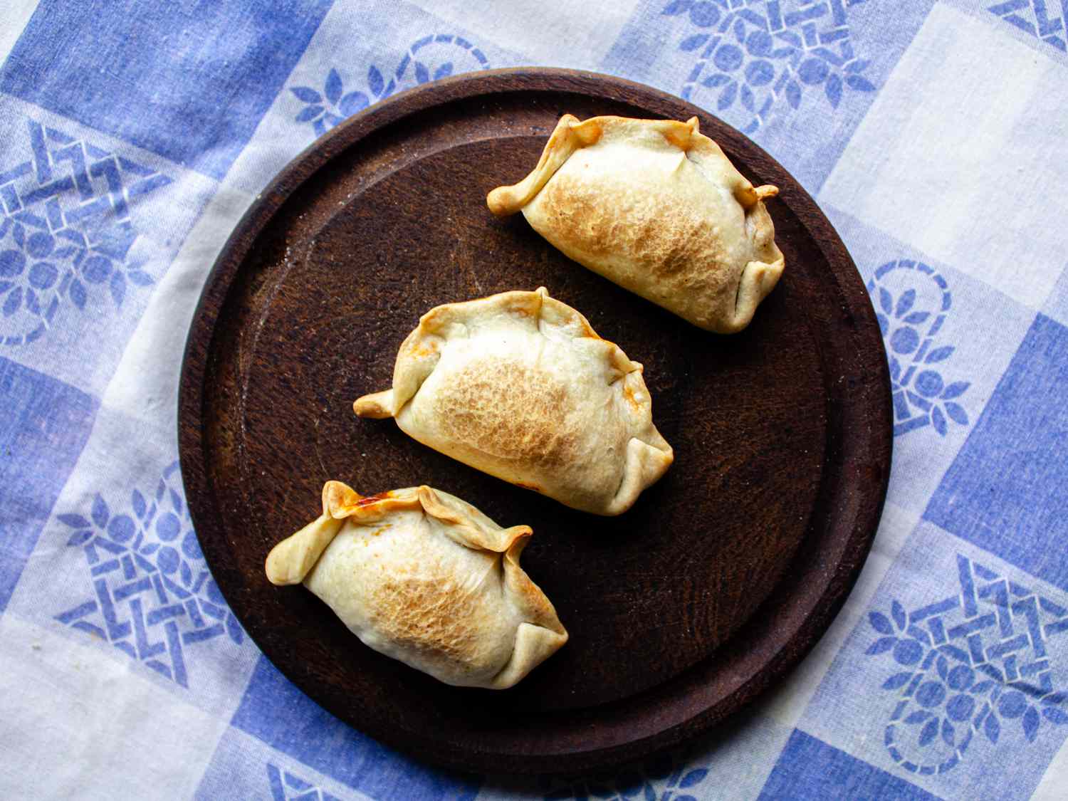 3 empanadas on a wooden plate on top of a traditional Argentinian fabric with a blue and white checkered pattern 