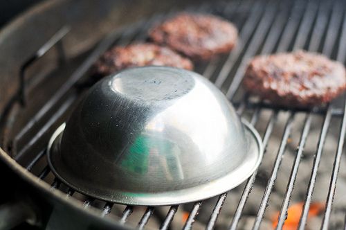 Three burgers on the grill, with a fourth underneath a stainless bowl to capture heat and finish cooking.