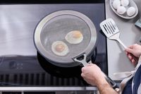 a person holding the le creuset splatter screen over a skillet with fried eggs.