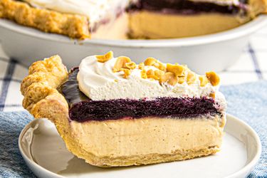 A slice of peanut butter and jelly pie topped with whipped cream and peanuts on a plate pie dish in the background