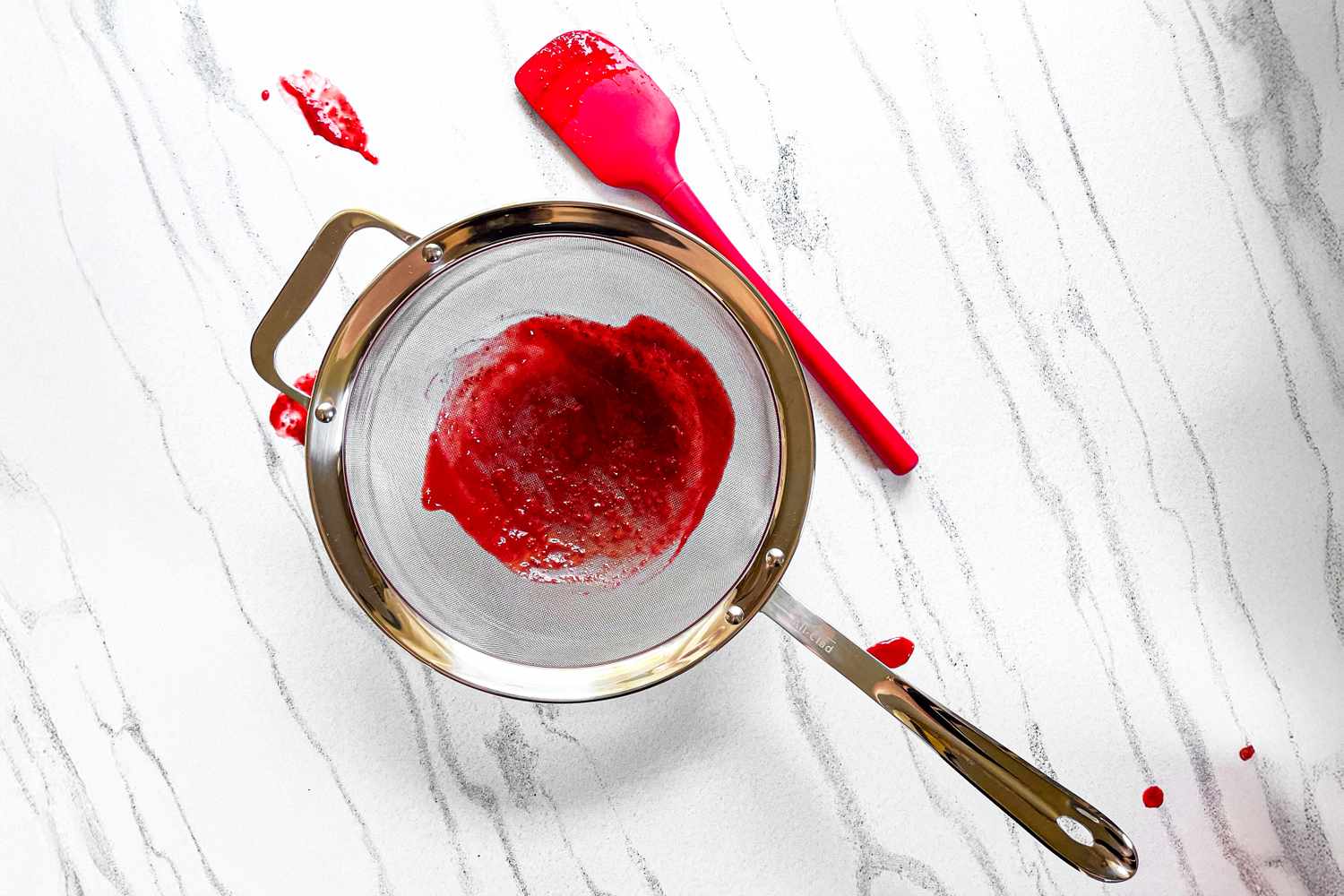 All-Clad Stainless-Steel Strainer, Set of 3 containing red puree placed on a marble surface with a red spatula next to it