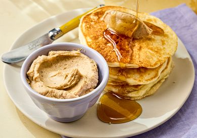 A plate of pancakes served with a small bowl of spreadable butter and honey with a yellowhandled knife nearby
