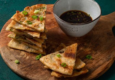 Scallion pancakes cut into slices, stacked on top of each other on a round wooden cutting board with a ceramic bowl of dipping sauce.