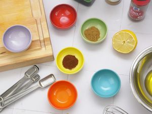 An assortment of small colorful bowls containing spices next to kitchen tools wooden board and a lemon half