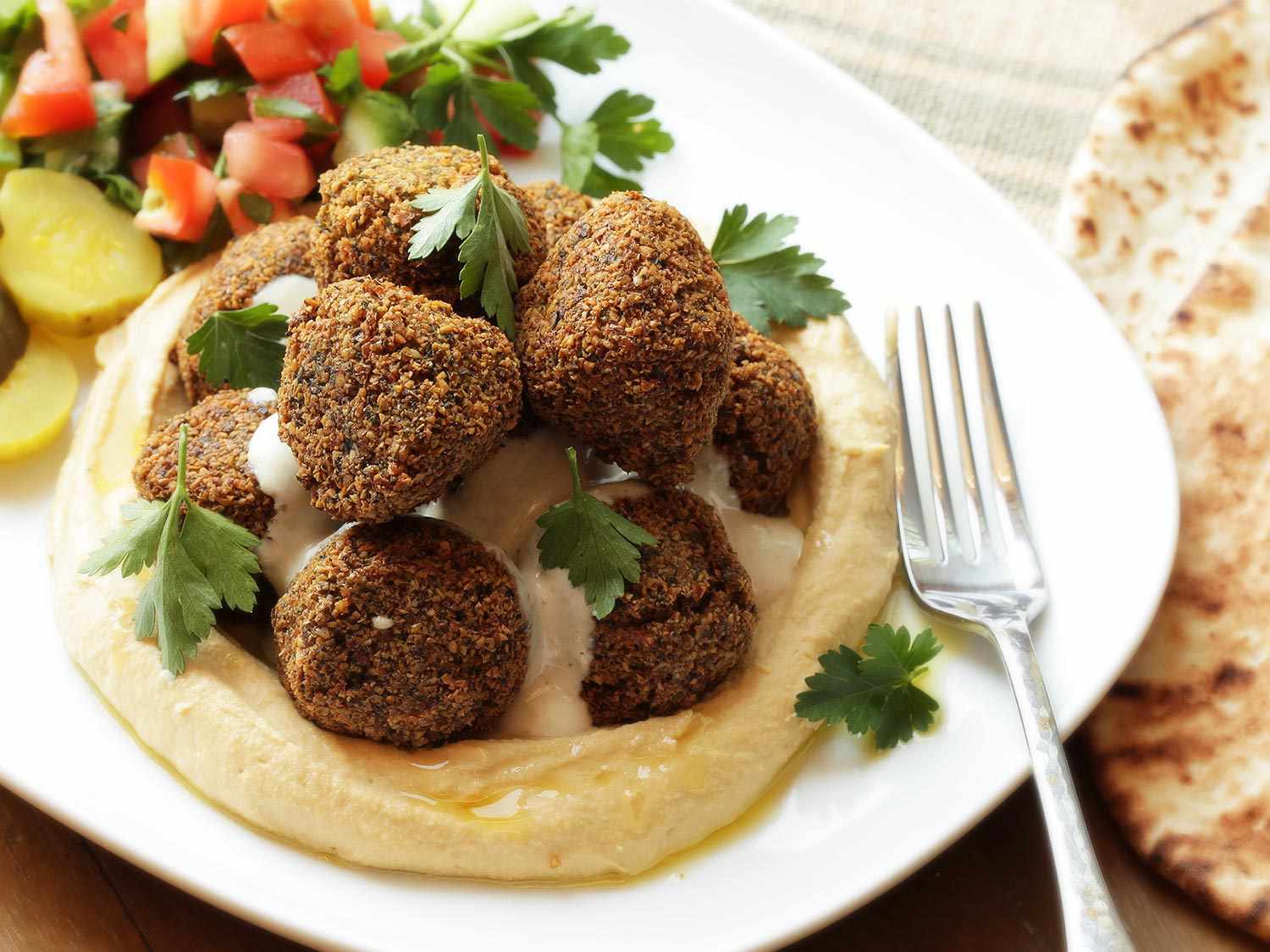 A plate showing multiple falafel balls piled on top of a round mound of hummus. There are also pickles and a chopped salad on the plate, and in the image are a metal fork and a pita bread.