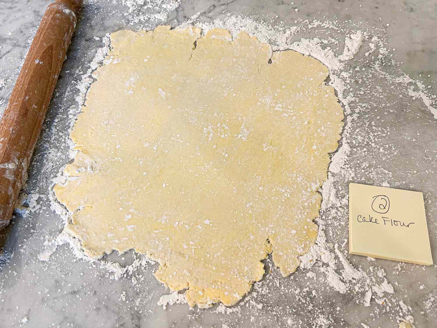 A sheet of rolled dough on a floured surface next to a wooden rolling pin and a labeled note card