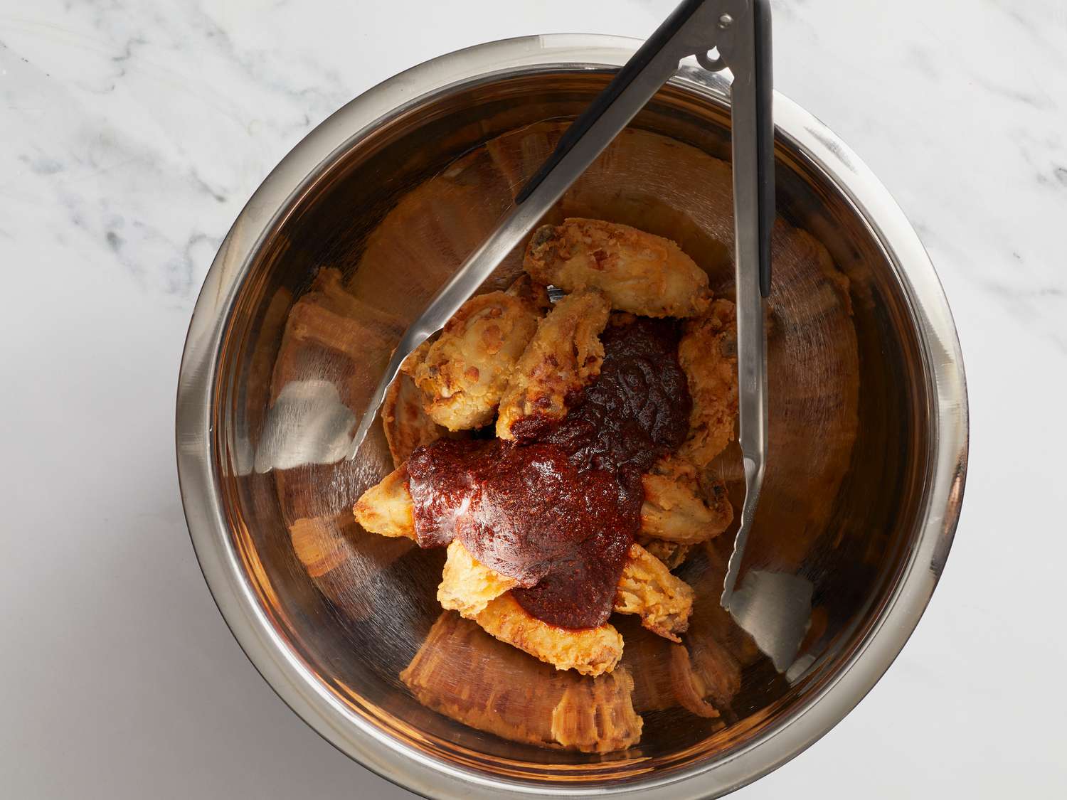 Pouring spice sauce on top of wings in bowl with tongs in it 