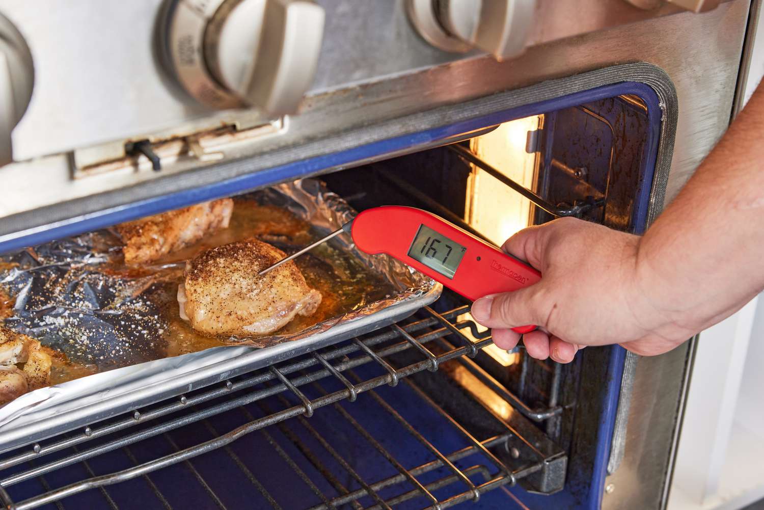 A hand using an instant-read thermometer to take the temperature of chicken thighs roasting in the oven