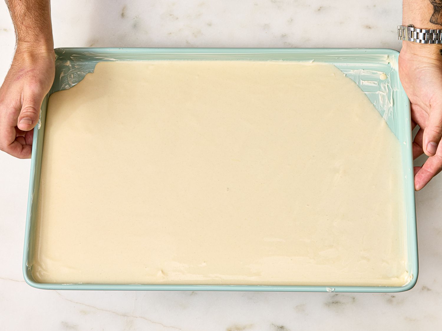 A person holding a rectangular tray filled with smooth batter on a marble surface