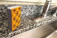 An orange Skura dishwashing sponge next to a kitchen sink
