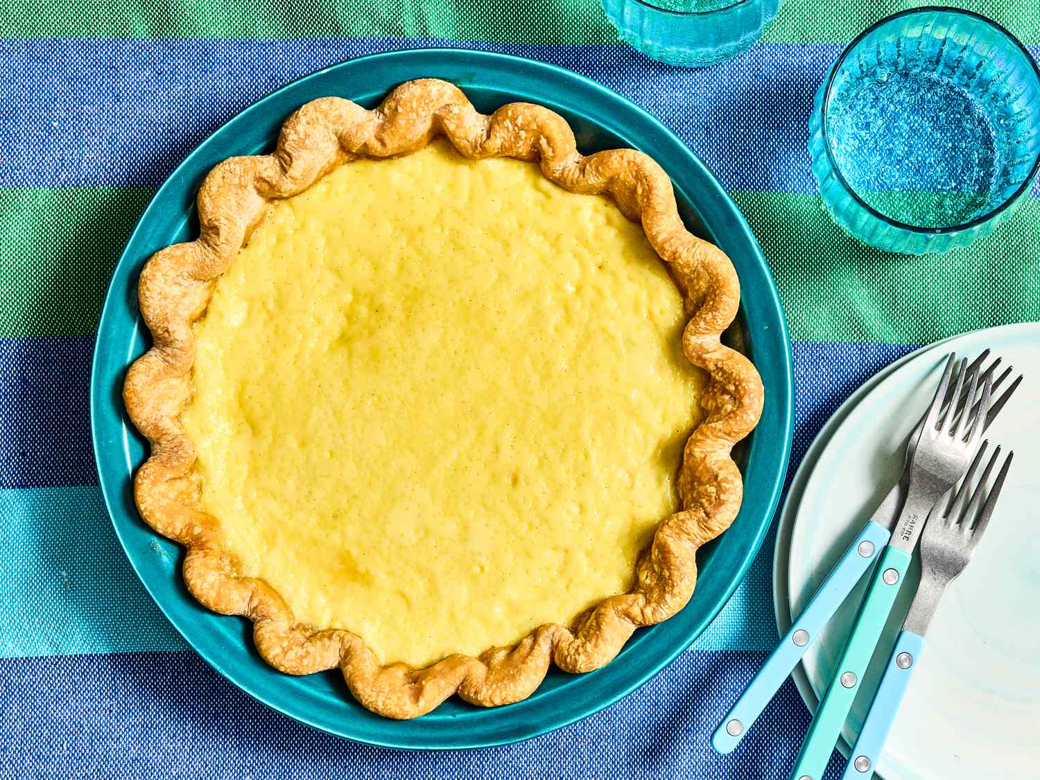 A custard pie garnished with a crimped crust, served on a turquoise plate with utensils and glassware on the side