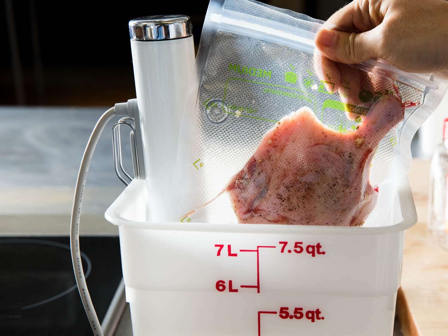 A hand lowering a vacuum-sealed bag of duck leg into a Cambro container with a sous vide circulator attached
