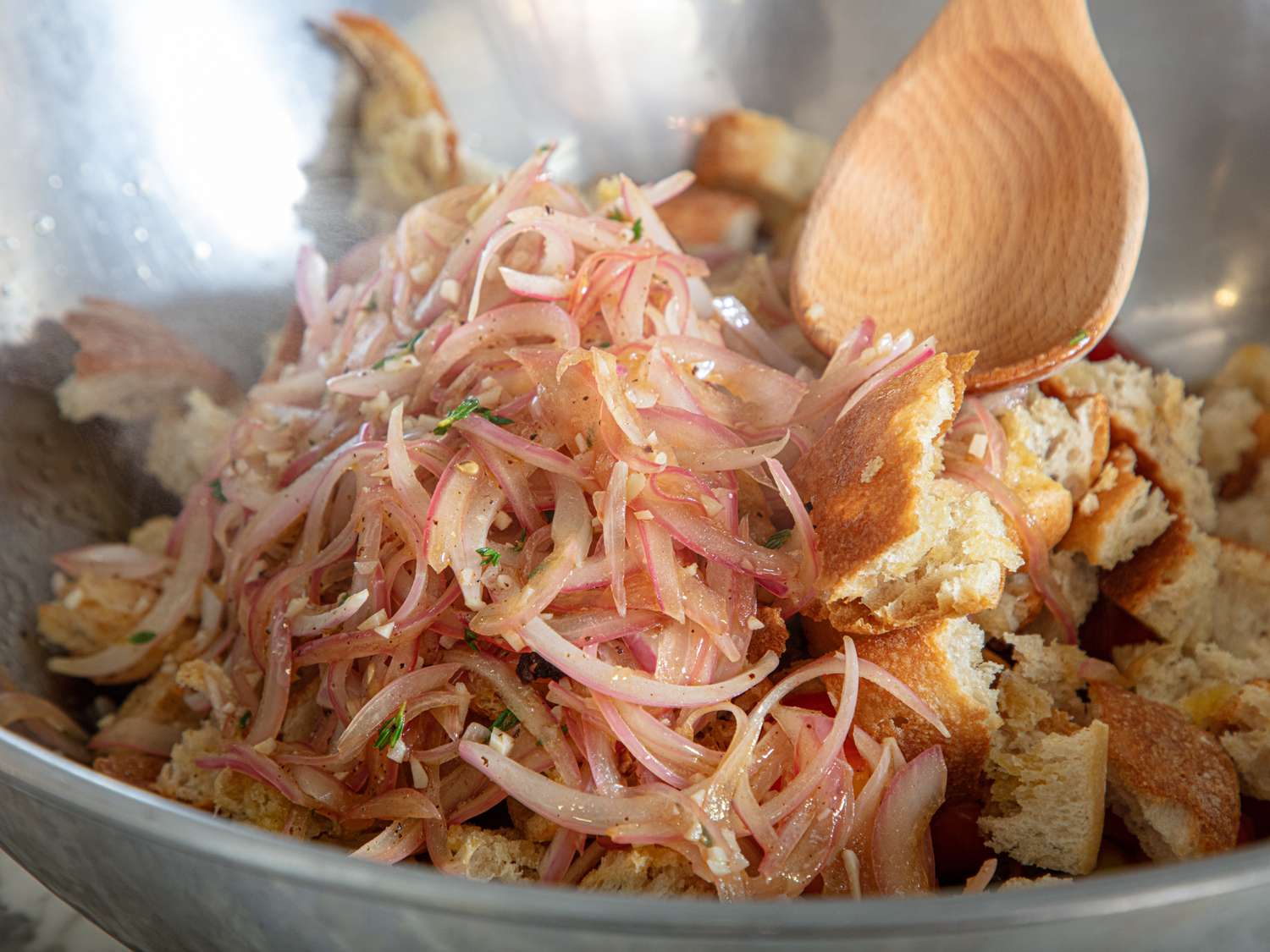 Mixture of torn bread pieces and sauted onions in a metal mixing bowl being stirred with a wooden spoon