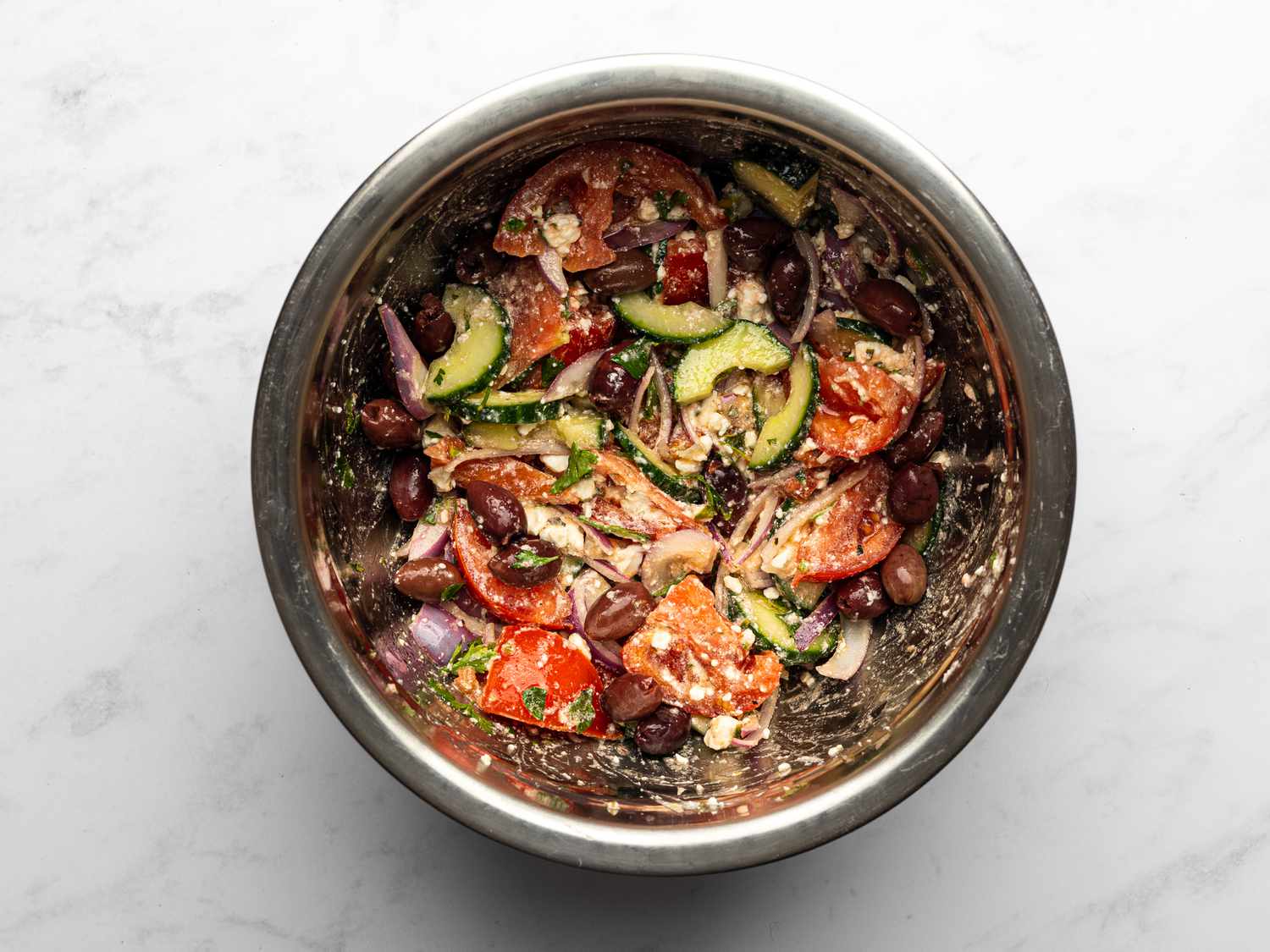 Olive oil whisked into reserved chicken marinade inside a bowl, with tomatoes, onions, cucumber, olives, feta, and parsley added, and seasoned with salt and pepper