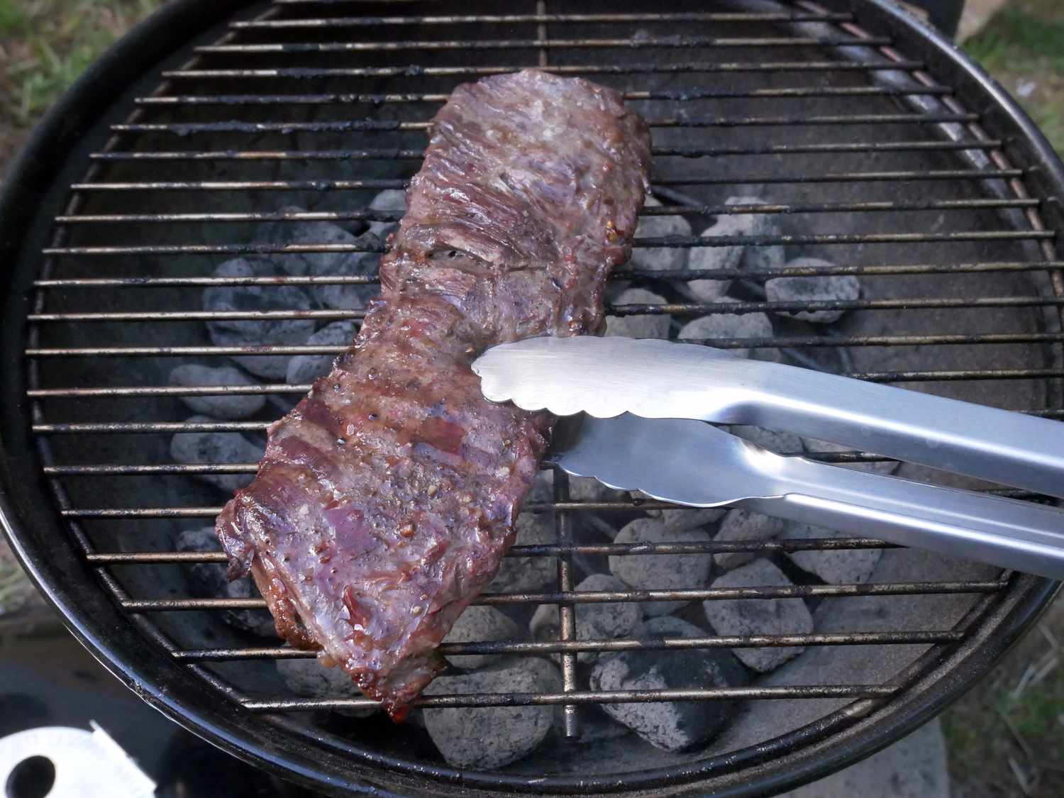 using tongs to grab a piece of flank steak on a charcoal grill