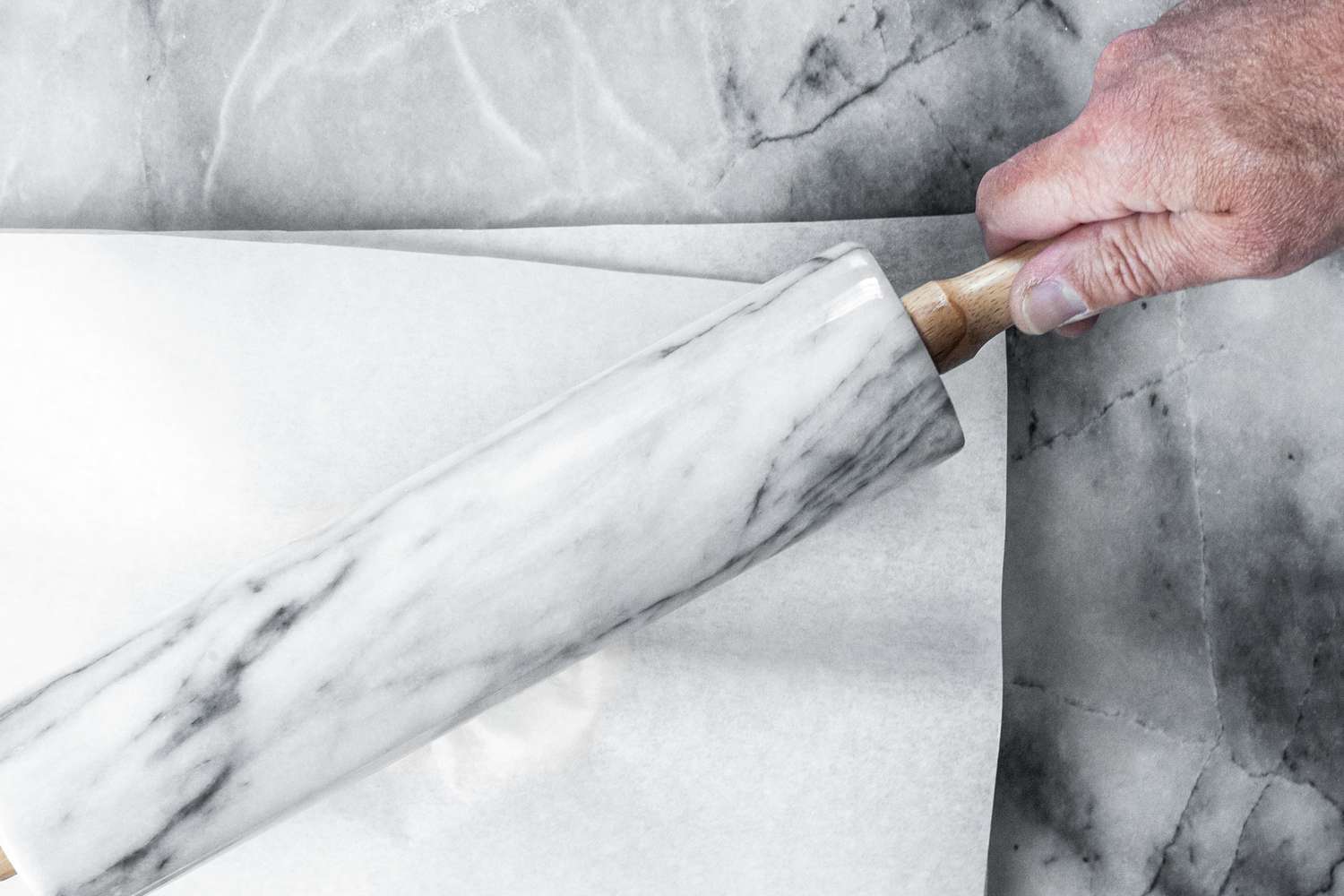 A hand holding a marble rolling pin above a sheet of parchment paper on a marble surface