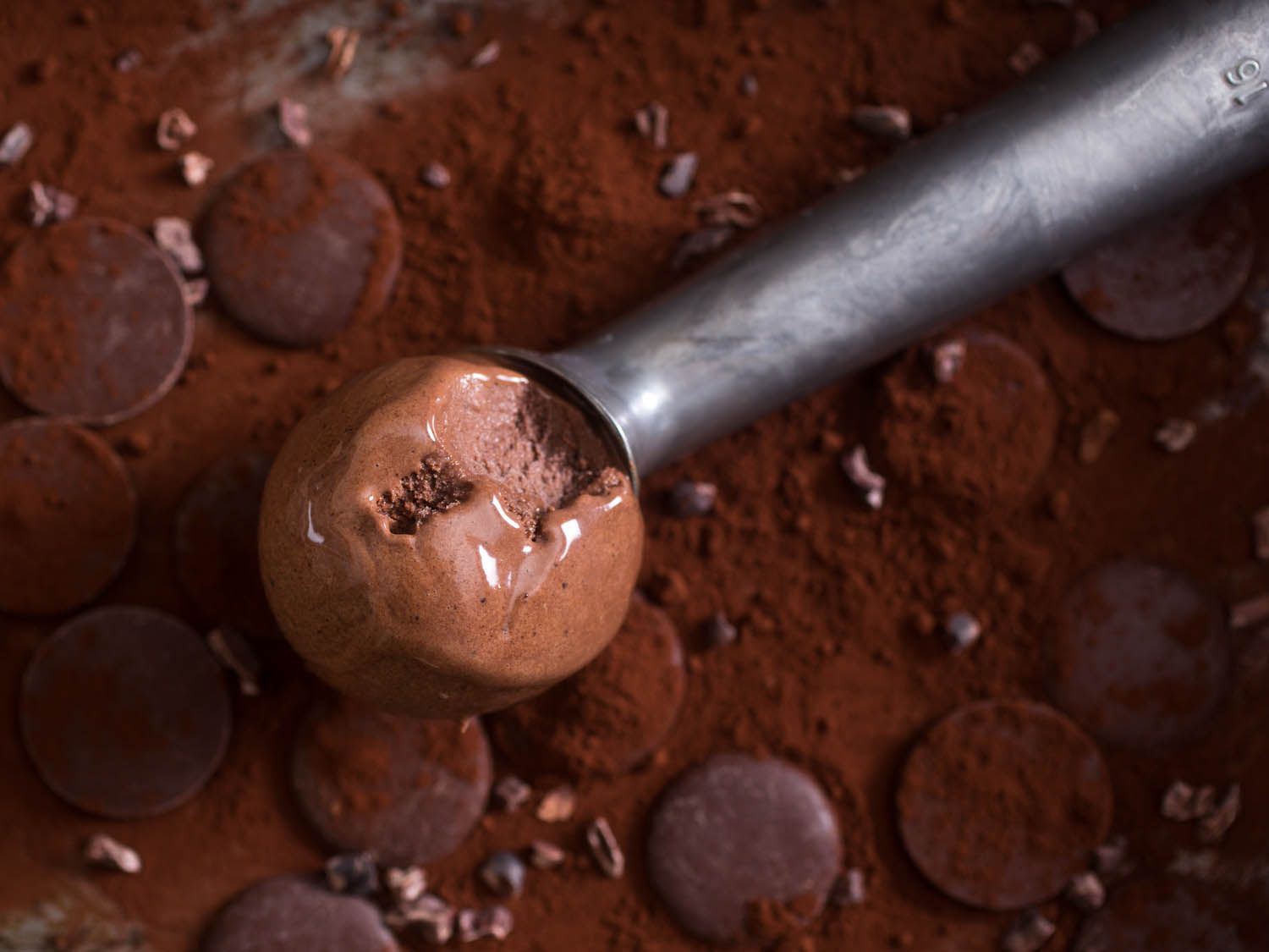 An ice cream scooper with a scoop of homemade chocolate ice cream. There are chocolate disks and cacao nibs in the background.