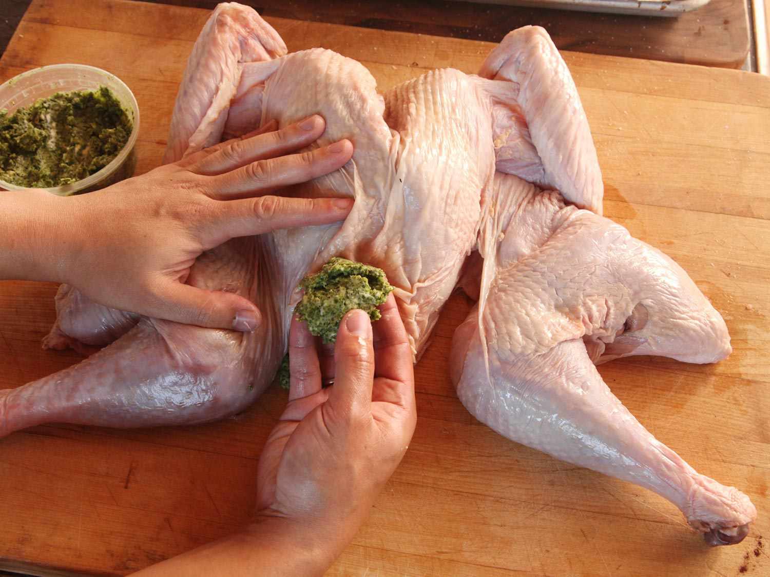 A hand holds an uncooked turkey steady while a second hand holds a large knob of herb butter, ready to be inserted underneath the skin.