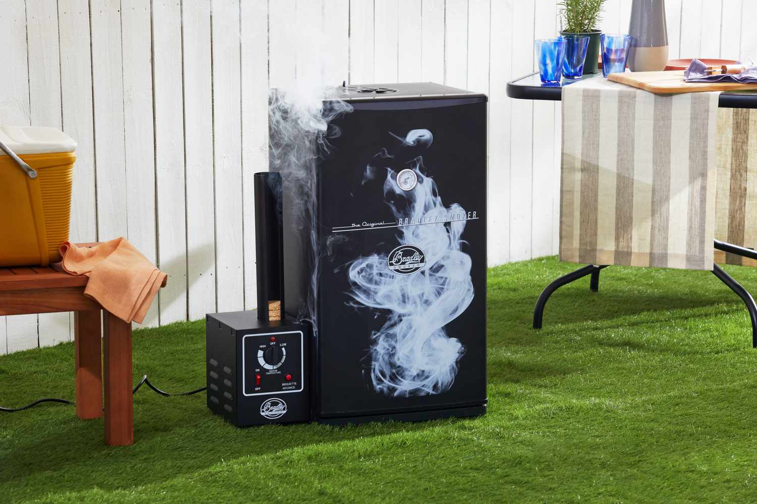 Electric smoker with visible smoke emitting outdoors, positioned near a white fence on grass