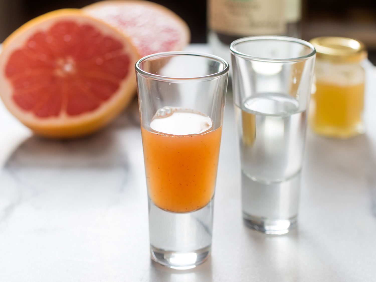 Closeup of a shot glass of grapefruit sangrita with chipotle and honey, flanked by another shot glass full of silver tequila. A halved grapefruit is in the blurred background.