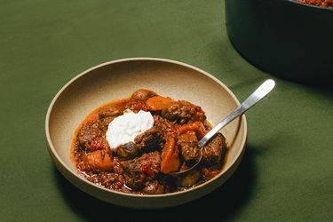 Bowl of beef stew with vegetables and sour cream served with a spoon pot of stew in the background