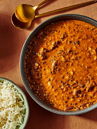 Dal Makhani in a bowl with 2 gold spoons, and 2 bowls of rice. Burnt orange colored surface and blue glass of water. 