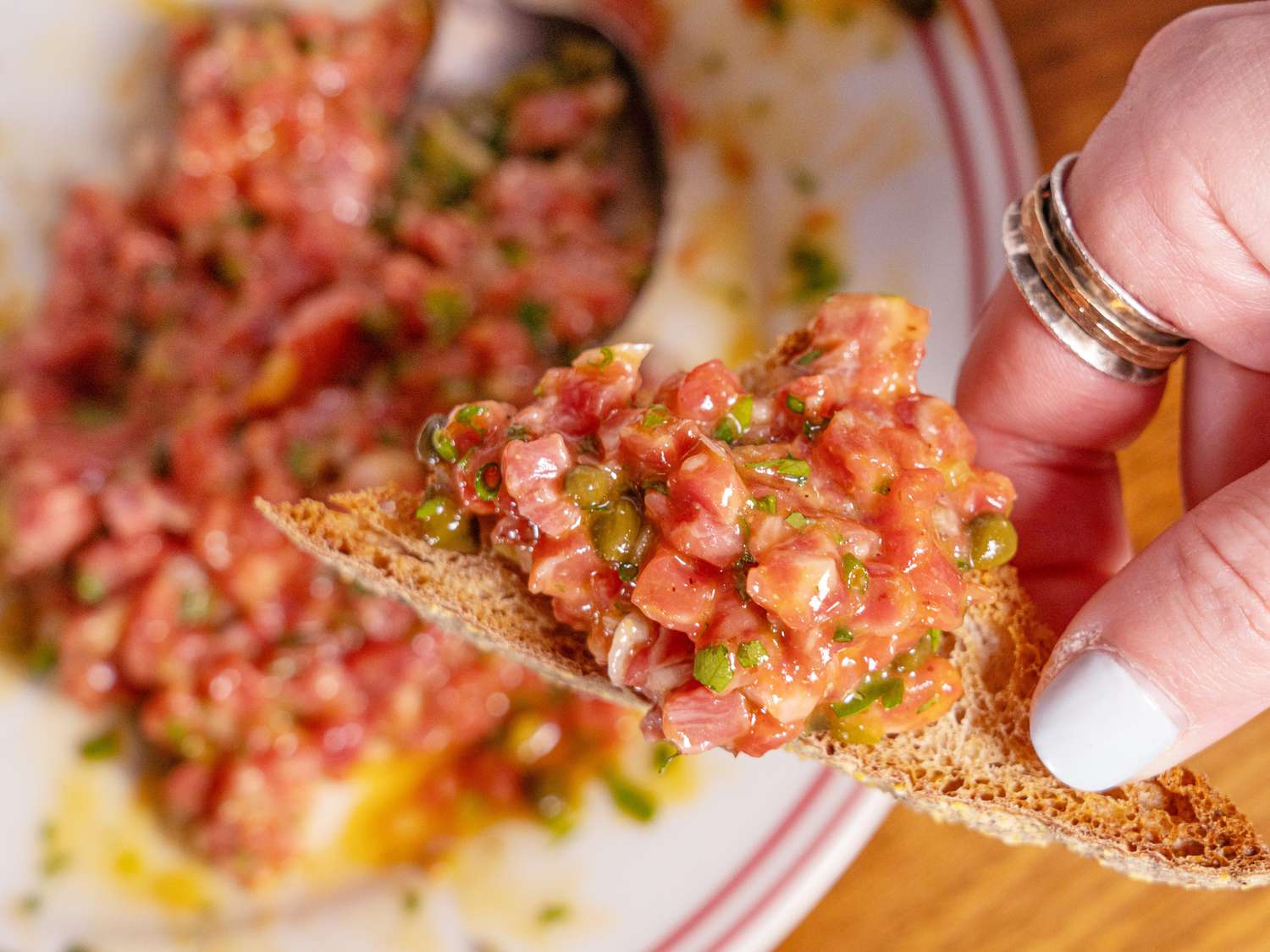 beef tartar spread on a small piece of bread, held above a dish of full tartar serving 