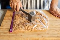 A person crushing cookies inside a sealed plastic bag using a meat tenderizer on a wooden cutting board