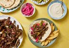 Beef shawarma wrap with accompaniments including pickles tahini sauce and pita bread on a table