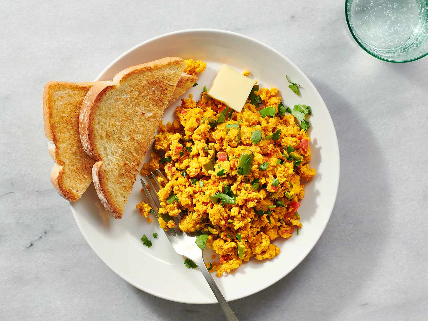The anda bhurji on a white ceramic plate with toasted white bread and a pat of butter on a stone counter 