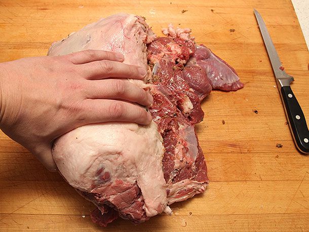 A hand rolling a boneless leg of lamb roast on a cutting board.