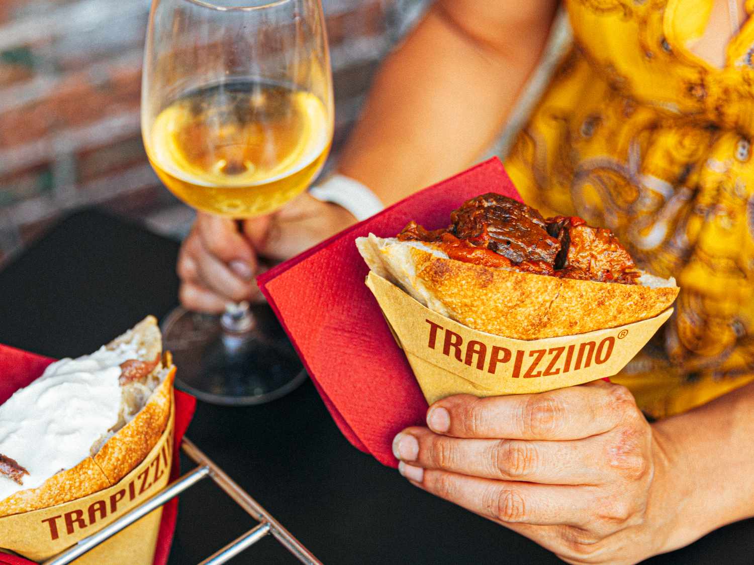A person in a yellow dress holding a trapizzino in one hand and a glass of wine in the other