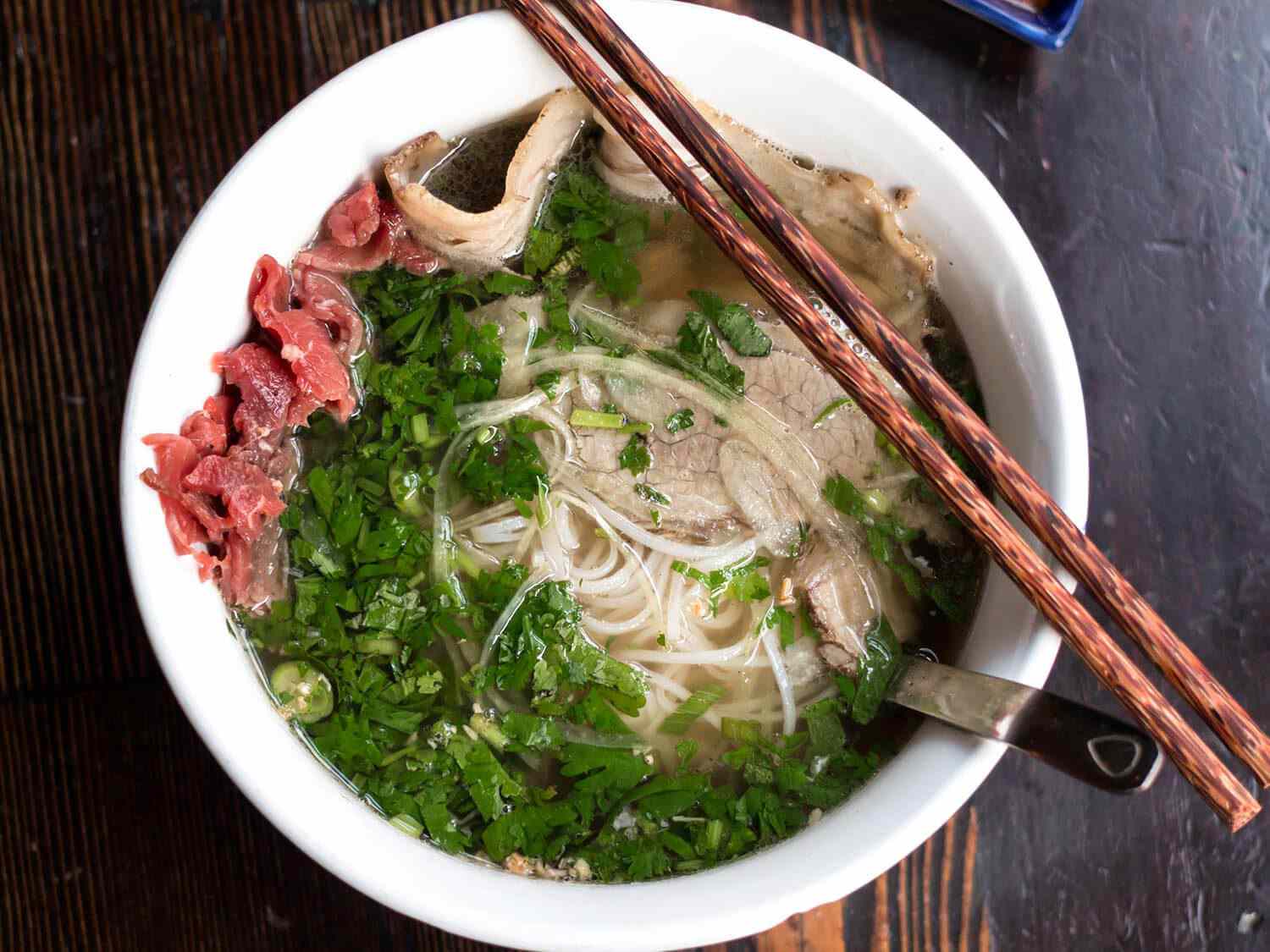 Overhead shot of bowl of pho with meat, noodles, herbs, and a pair of chopsticks resting on the side of the bowl