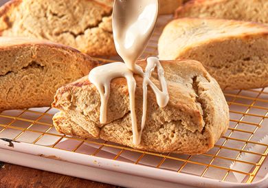 Maple scone being drizzled with glaze displayed on a cooling rack with other scones in the background