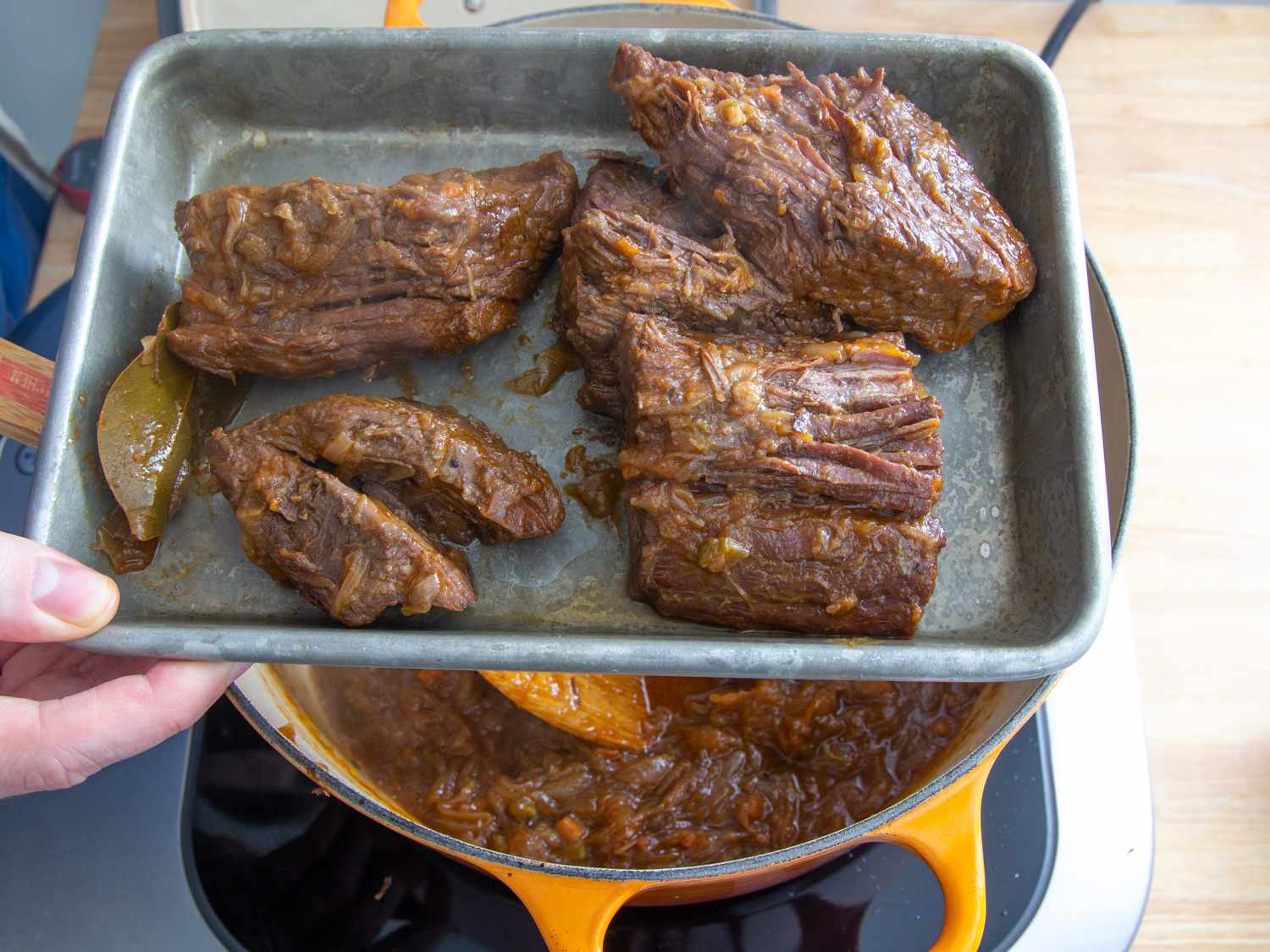 A baking pan holding the cooked pieces of beef and bay leaf above a Dutch oven with cooked vegetables. 