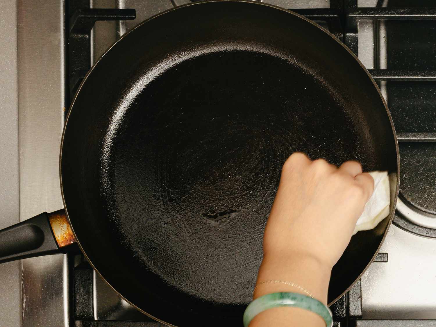 A hand wiping a cast iron skillet on a stove with a cloth