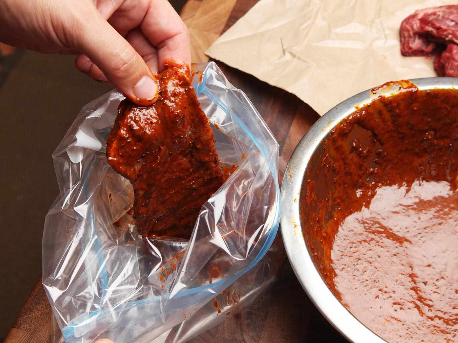 A hand adding a skirt steak that has been dipped in carne asada marinade to a large zipper lock bag. A bowl of marinade and un-coated steaks are in the background.