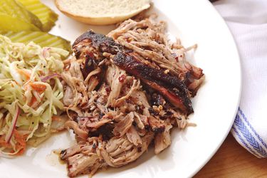 A plate of sous vide pulled pork with slaw, pickle slices, and a bun.