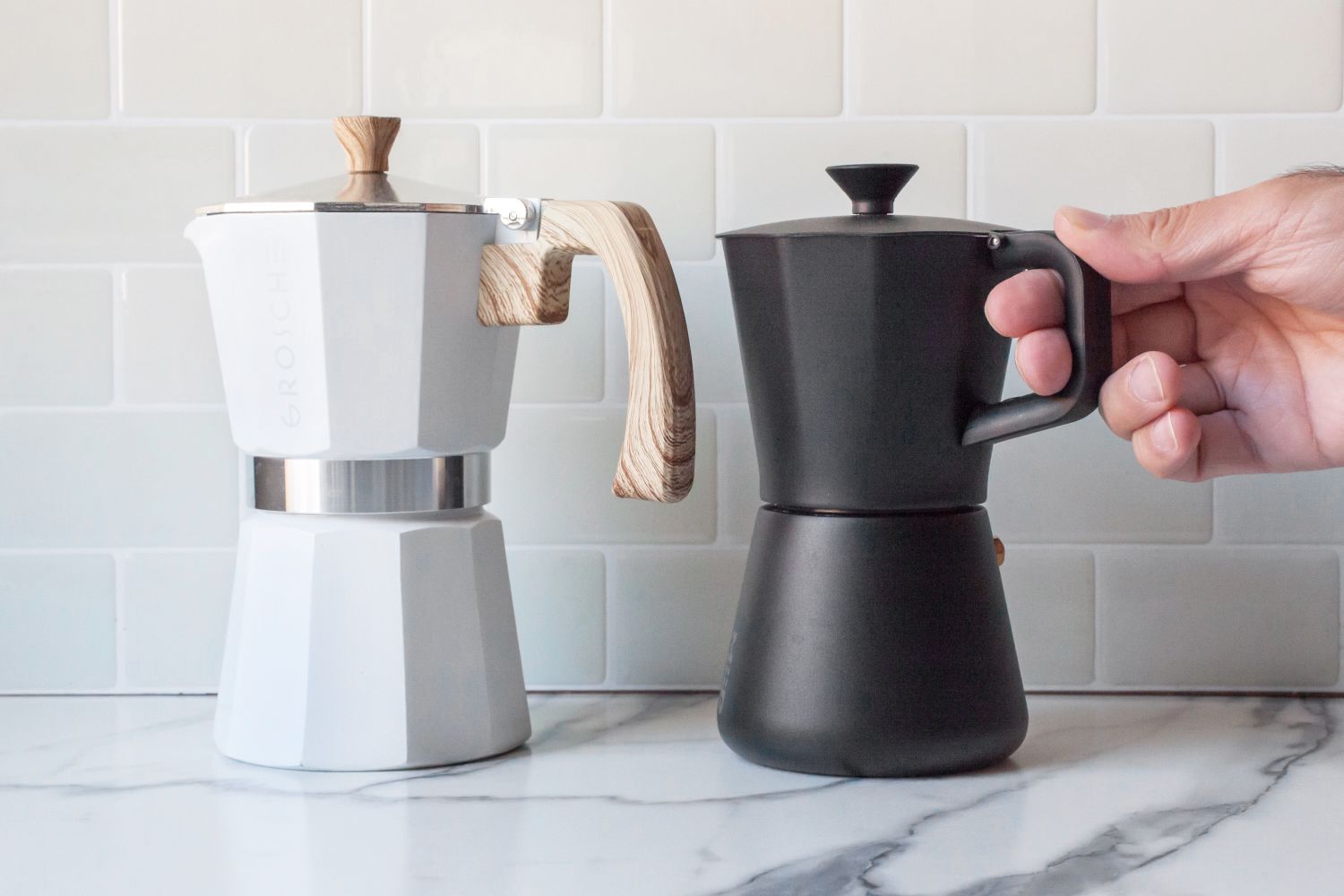 A person holding onto the small handle of a moka pot next to a moka pot with a larger handle