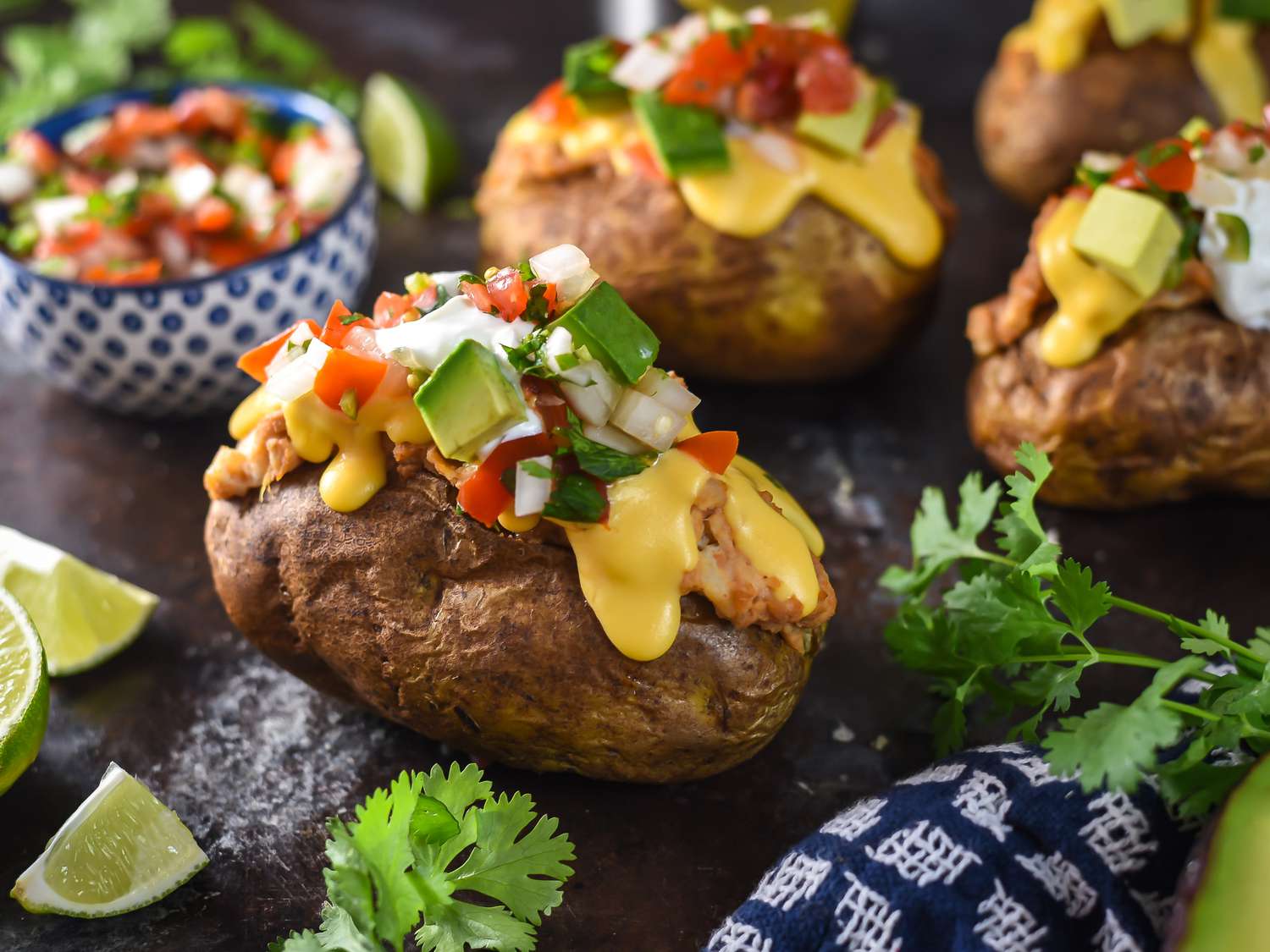 Several loaded backed potatoes, nacho-style, with cheese sauce, pico de gallo, sour cream, avocado, and beans. Several potatoes are pictured along with fresh cilantro, lime wedges, and a bowl of pico.