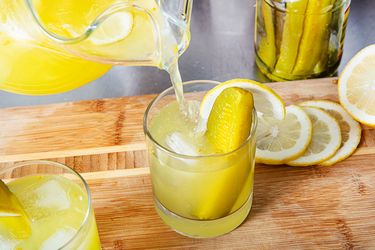Glass of lemonade with a pickle spear lemon slices and a mason jar with pickles nearby on a wooden surface