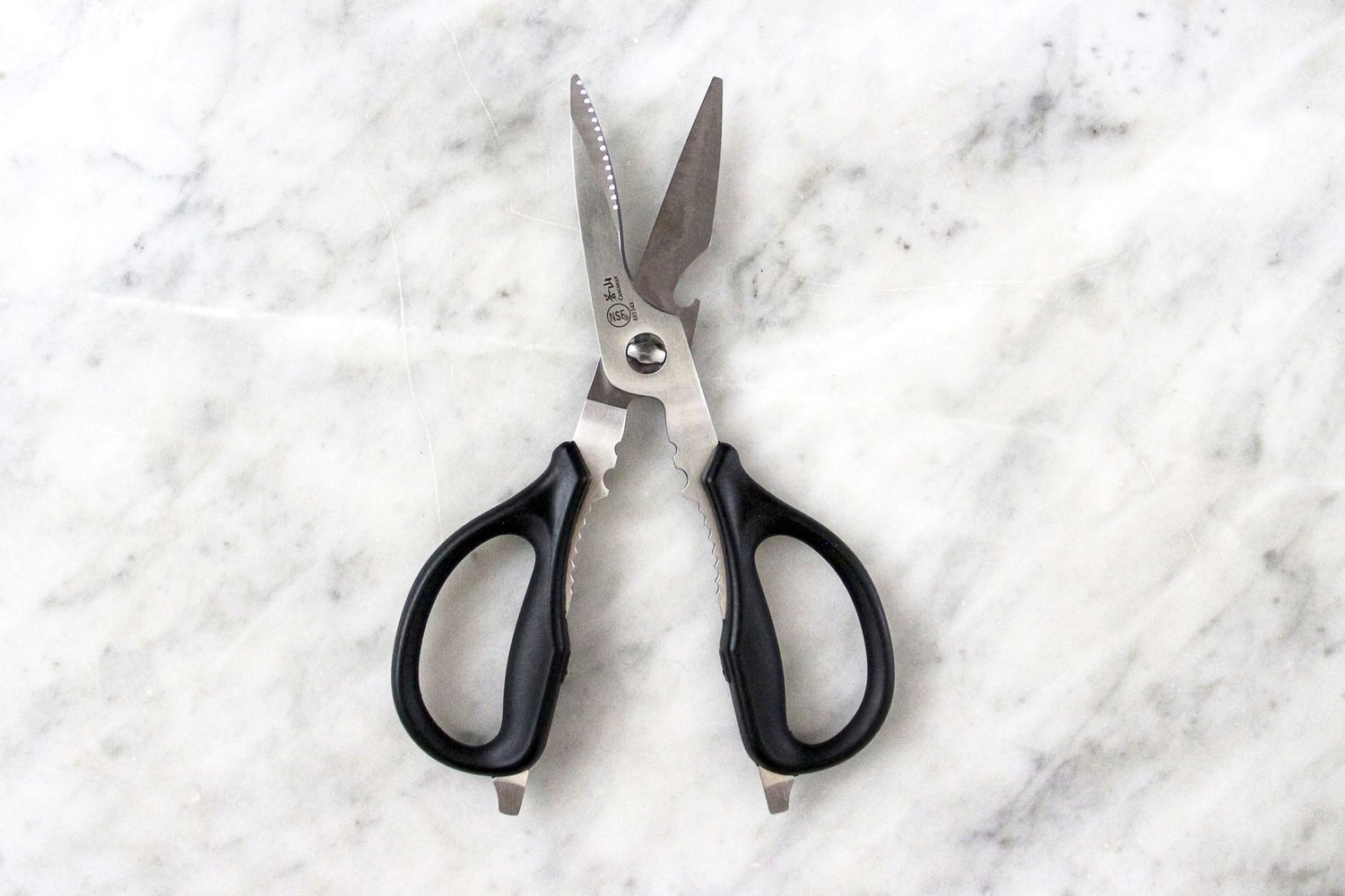 A pair of Cangshan 9-inch Heavy-Duty Come-Apart Kitchen Shears with black handles and serrated blades placed on a marble surface