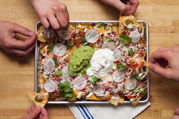 Hands grabbing chips from a tray of fully-loaded nachos
