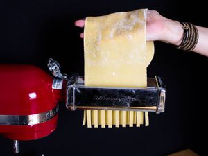 a hand feeding a sheet of pasta through a stand mixer's pasta cutter attachment