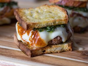 Three asado burgers on a wood cutting board, each made on sliced bread and topped with melted cheese and chimichurri. 