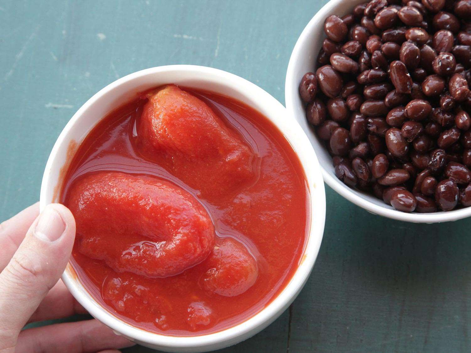 Hand holding a bowl filled with canned roma tomatoes and a bowl filled with black beans